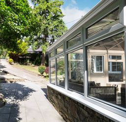 A paved pathway beside a conservatory with glass windows and garden with trees and shrubs at Nant Cottage in Rowen