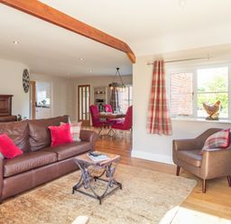 A living room with a brown leather sofa and armchair red and plaid cushions beside a dining area with red chairs and a window with plaid curtains at The Croft in Thompson near Watton