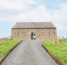 A gravel driveway leading to a stone building with a slate roof and an arched door surrounded by green grass at Gwel Y Fenai in Y Felinheli