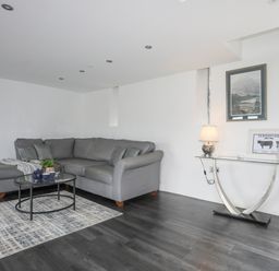 A living room with a grey sectional sofa a glass coffee table with a teapot and cups a patterned rug a glass side table with a lamp framed pictures and a plant at Gwel Y Fenai in Y Felinheli