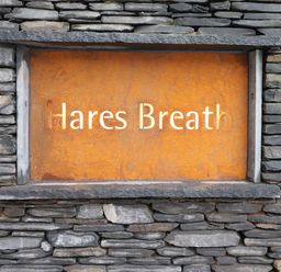 A rust-colored metal sign with the text Hares Breath mounted on a stone wall at Hares Breath in Bowness-On-Windermere