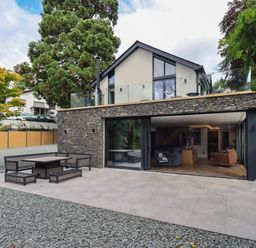 An outdoor seating area with table and benches on a tiled patio in front of a modern two-story house with stone and glass exterior at Hare's Breath in Bowness-On-Windermere