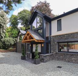 The exterior of a modern house with a stone facade large windows a wooden entrance canopy and gravel driveway surrounded by trees at Hares Breath in Bowness-On-Windermere
