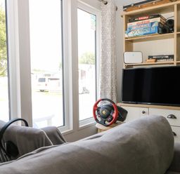 A living room with a sofa a television a gaming steering wheel and board games on shelves at The Goodwood Lodge in Runcton near Chichester