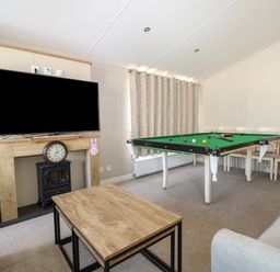 A living room with a television above a fireplace, a clock, a pool table, and chairs at The Goodwood Lodge in Runcton near Chichester