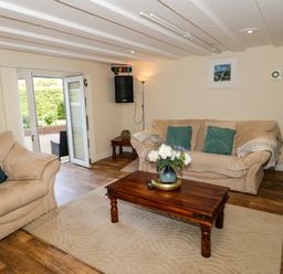 A living room with beige sofas green cushions a wooden coffee table with flowers hardwood floor and open doors to a garden at Plas Eithin in Mynydd Mechell near Llanfechell