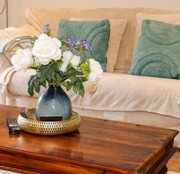A wooden coffee table with a blue vase of white and purple flowers a gold tray and a remote control in front of a cream sofa with teal cushions and a throw at Plas Eithin in Mynydd Mechell near Llanfechell