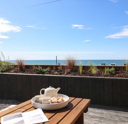A wooden table with a teapot cups and pastries with a view of the sea and plants at Sandpiper Chalet in Eype