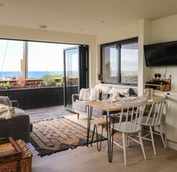 A living room with sofas and a dining table near an open door overlooking the sea at Sandpiper Chalet in Eype
