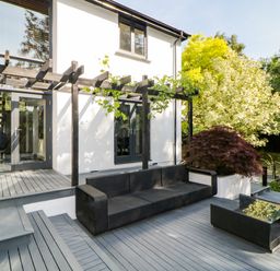 An outdoor patio with black wicker sofa and table next to a white house with wooden pergola and plants at Iroko in Millbrook