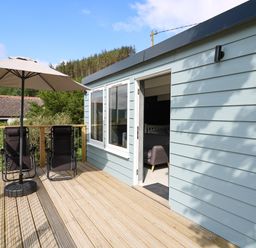 A wooden deck with two black chairs under a beige umbrella outside a light blue building at Caban Glas in Llanafan near Pontrhydfendigaid