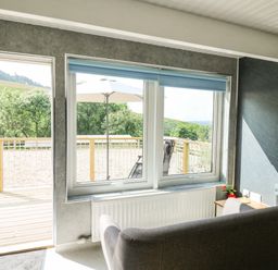 A room with a gray sofa facing windows and a door leading to a wooden deck with an umbrella and chairs at Caban Glas in Llanafan near Pontrhydfendigaid
