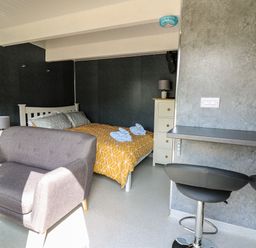 A bedroom with a bed with yellow and gray bedding a gray sofa a floor lamp a chest of drawers and two black bar stools at Caban Glas in Llanafan near Pontrhydfendigaid