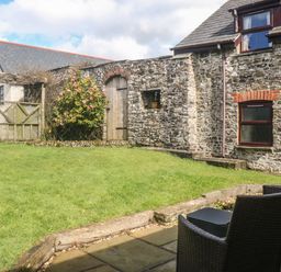 A garden with grass a stone wall and wooden door next to a stone house with wicker chairs on a patio at Honey Bee Cottage in Bradworthy