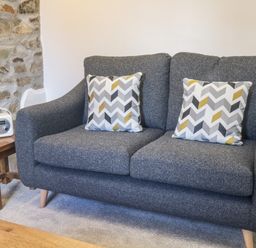 A grey sofa with two patterned cushions next to a wooden side table with a small radio on it at Honey Bee Cottage in Bradworthy