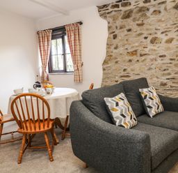 A dining area with a round table and wooden chairs next to a dark gray sofa with patterned cushions by a stone wall at Honey Bee Cottage in Bradworthy