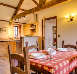 A dining area with a table set for two near a kitchen with wooden cabinets and exposed wooden beams at Woodpecker Cottage in Bradworthy near Bude