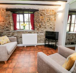 A living room with stone walls a window with red curtains two sofas with yellow pillows a television on a stand and a floor lamp at Wagtail in Bradworthy near Bude