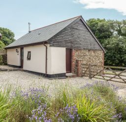 A small detached house with a gravel driveway and a wooden gate surrounded by greenery and flowers at Jenny Wren in Bradworthy