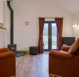 A living room with two armchairs a wood burning stove and glass doors with curtains at Jenny Wren in Bradworthy