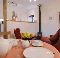 A dining table with white teapot cups and a bowl of fruit near red armchairs and a wood stove in a kitchen and living room at Jenny Wren in Bradworthy