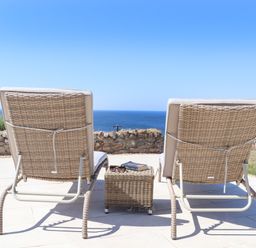 Two wicker lounge chairs and a wicker side table on a stone patio overlooking the ocean at Boscregan in Porthleven