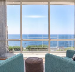 Two blue armchairs and a brown ottoman facing large windows overlooking the sea at Boscregan in Porthleven