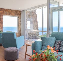 A living room with blue armchairs a sofa flowers a stone wall and ocean view at Boscregan in Porthleven