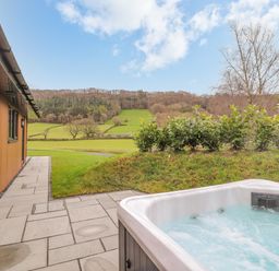 An outdoor hot tub on a stone patio next to a wooden building overlooking grassy hills and trees at Pool View in Meifod Powys