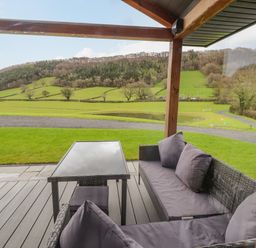 A covered outdoor patio with a glass table and wicker seating cushions overlooking fields and hills at Pool View in Meifod Powys
