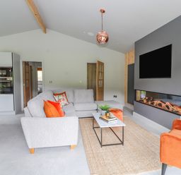 A living room with a grey sectional sofa with orange and floral cushions a rectangular coffee table and an orange armchair next to a modern fireplace and mounted TV at Pool View in Meifod Powys