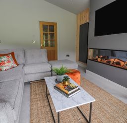 A living room with a gray sectional sofa with floral and orange cushions a marble coffee table with books and plants a wall-mounted TV and an electric fireplace at Pool View in Meifod Powys