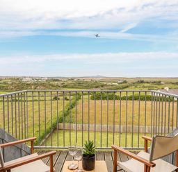 A balcony with two chairs a small table with two glasses and a plant overlooking fields and distant houses at Plas Newydd in Llanfaelog near Rhosneigr