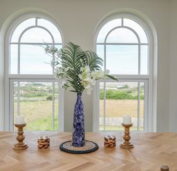 A wooden table with a blue vase of flowers two wooden candle holders and two small wooden boxes in front of arched windows at Plas Newydd Llanfaelog near Rhosneigr