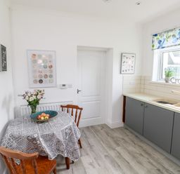 A kitchen with a round table covered with a patterned tablecloth two wooden chairs a bowl of apples flowers on the table grey cabinets and a window with a floral blind at Garth in Pen-y-bryn near Cardigan