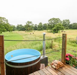 An outdoor wooden hot tub on a deck with firewood and flowers overlooking a field with trees at The Lazy Mare by The Water Admaston near Abbots Bromley