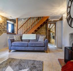 A living room with a sofa clock wood stove staircase and stone walls at Hollytree Cottage in Hook Norton