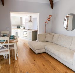 An open living and dining area with a beige sofa a wooden dining table set with plates and bowls and a kitchen with white cabinets and appliances at Ramsey Cottage in Milford Haven