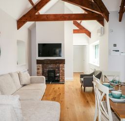 A living room with a beige sofa a wall-mounted TV above a brick fireplace wooden ceiling beams and a dining table set with green dishes at Ramsey Cottage in Milford Haven