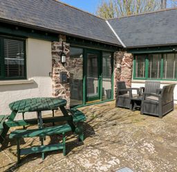 An outdoor patio with a green wooden picnic table and four benches and two gray single chairs with a small table outside a stone and white plaster building with green framed windows and door at Ramsey Cottage in Milford Haven