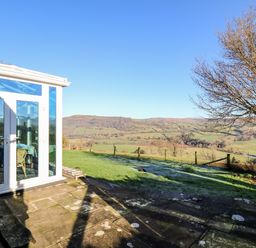 A patio area next to a conservatory with a view of fields hills and a tree at Ty Twmp Tump Cottage in Tal-Y-Bont-On-Usk