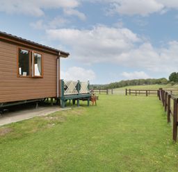 A side view of a brown wooden cabin with a small porch and green railings next to a fenced grassy field with trees in the distance at Hill View Lilac Lodge in Wareham