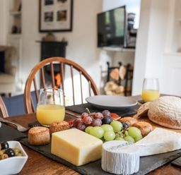 A dining table with cheese, grapes, bread, olives, and glasses of orange juice in a room with wooden chairs and a fireplace at Silva in Penmon