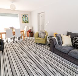 A living room with a striped carpet a dining table with chairs a green armchair a grey sofa with cushions and a painting on the wall at Seagull Cottage in Bude