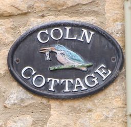 A wall-mounted oval plaque with a bird holding a fish and text reading Coln Cottage on a stone wall at Coln Cottage in Stow-On-The-Wold