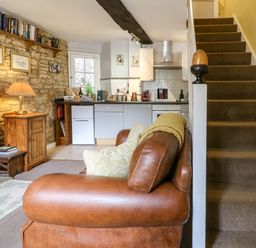 A living area with a brown leather sofa next to carpeted stairs and a kitchen with light gray cabinets at Coln Cottage in Stow-On-The-Wold