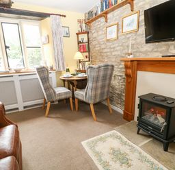 A sitting area with two checkered chairs at a small round table near a window and an electric stove under a mounted television at Coln Cottage in Stow On The Wold