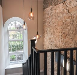 A stairway with black railing next to a stone wall and arched window with hanging pendant lights at The Sigma Penthouse in Dyserth
