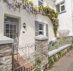 The exterior of a white house with a gray door and window shutters a small garden with purple flowers and a stone and metal fence at Saltwater Cottage in Babbacombe