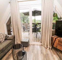 A living room with a sofa, two black metal wire tables, a television on a wooden stand, sliding glass doors with curtains leading to a balcony with outdoor furniture and an umbrella at Lavender Lodge in St Ann's Chapel Cornwall